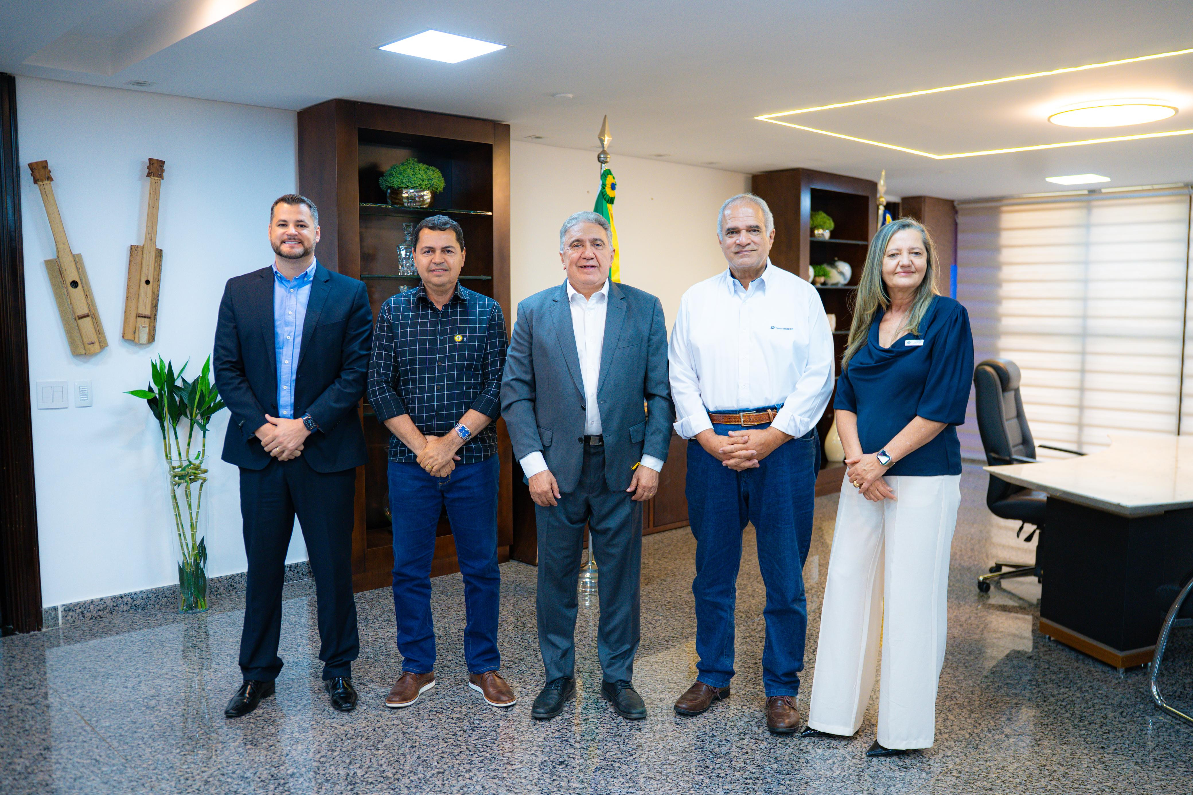 A imagem mostra cinco pessoas, todas de pé e sorrindo. Há três homens mais velhos, um homem mais jovem e uma mulher. Ao fundo, há uma estante com livros e a bandeira do Brasil. O ambiente é formal.