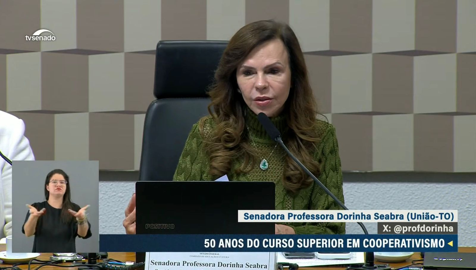 A imagem mostra uma mulher jovem falando em um microfone. Ela usa um suéter verde e está sentada em frente a um laptop. No canto da imagem, há uma pequena tela com uma intérprete de libras (língua de sinais). A legenda indica que a mulher é a senadora Professora Dorinha Seabra, falando sobre os 50 anos do curso superior em cooperativismo. Ao fundo, há uma parede com um padrão geométrico discreto.
