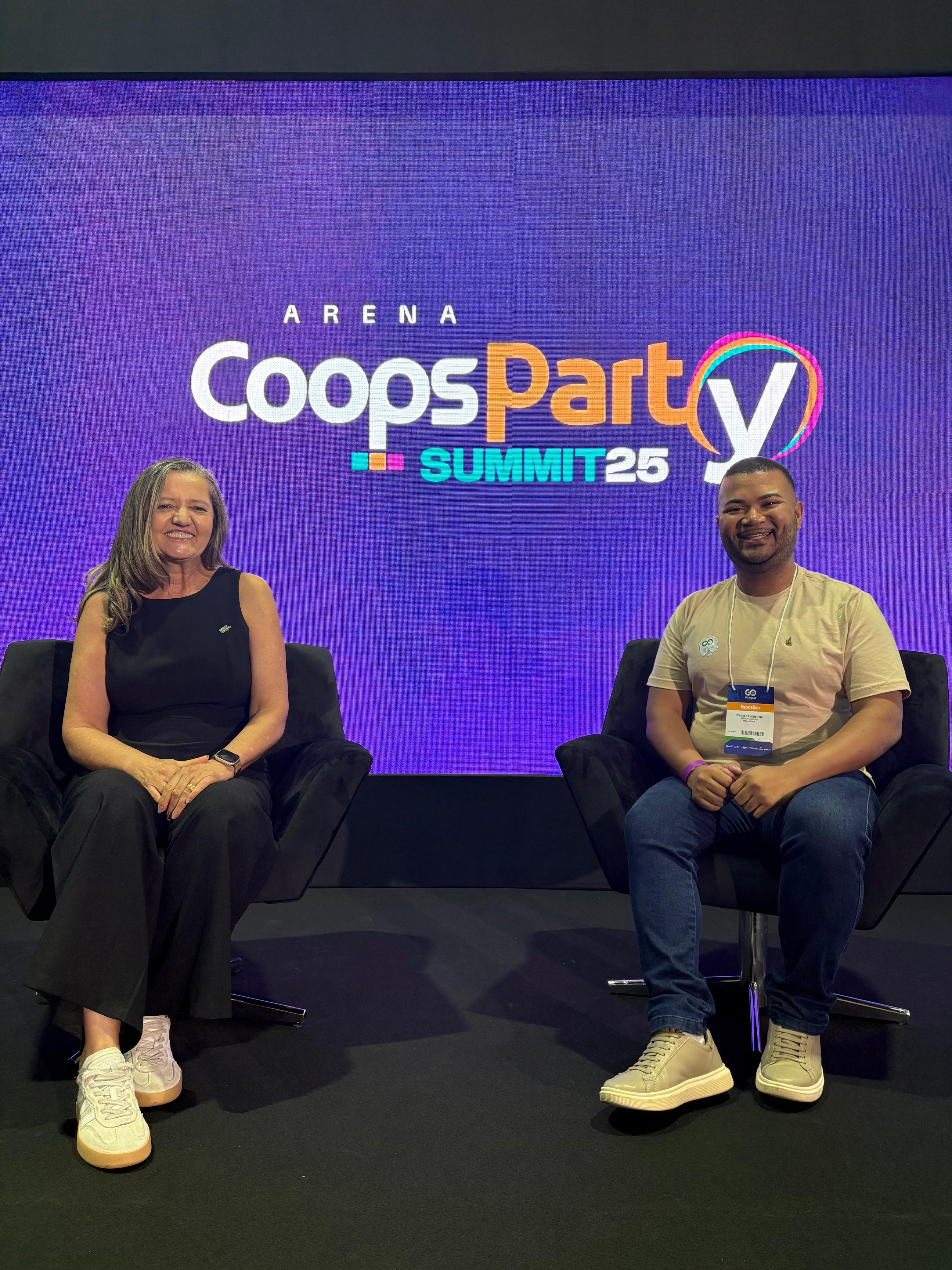A imagem mostra duas pessoas sentadas em poltronas pretas, diante de um painel roxo com o logotipo “Arena CoopsParty Summit 25”. Ambas sorriem para a câmera, em um ambiente de evento corporativo voltado à inovação e ao cooperativismo.