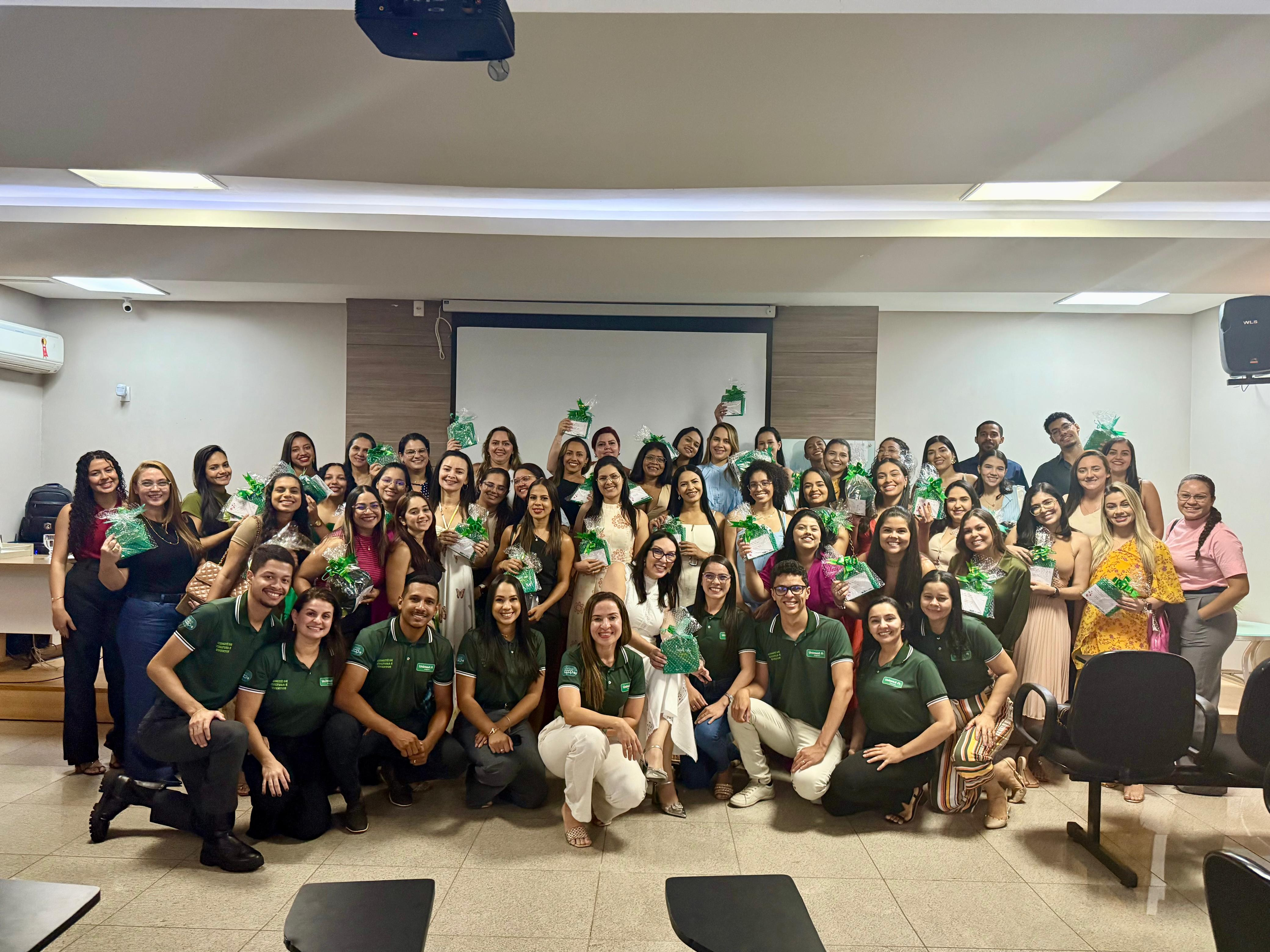 Esta é uma foto em grupo de muitas mulheres, tirada em uma sala com luzes no teto e uma tela de projeção ao fundo. Elas estão sorrindo e segurando pequenos presentes verdes. Algumas estão em pé, outras agachadas na frente, todas próximas umas das outras, mostrando um momento de confraternização.