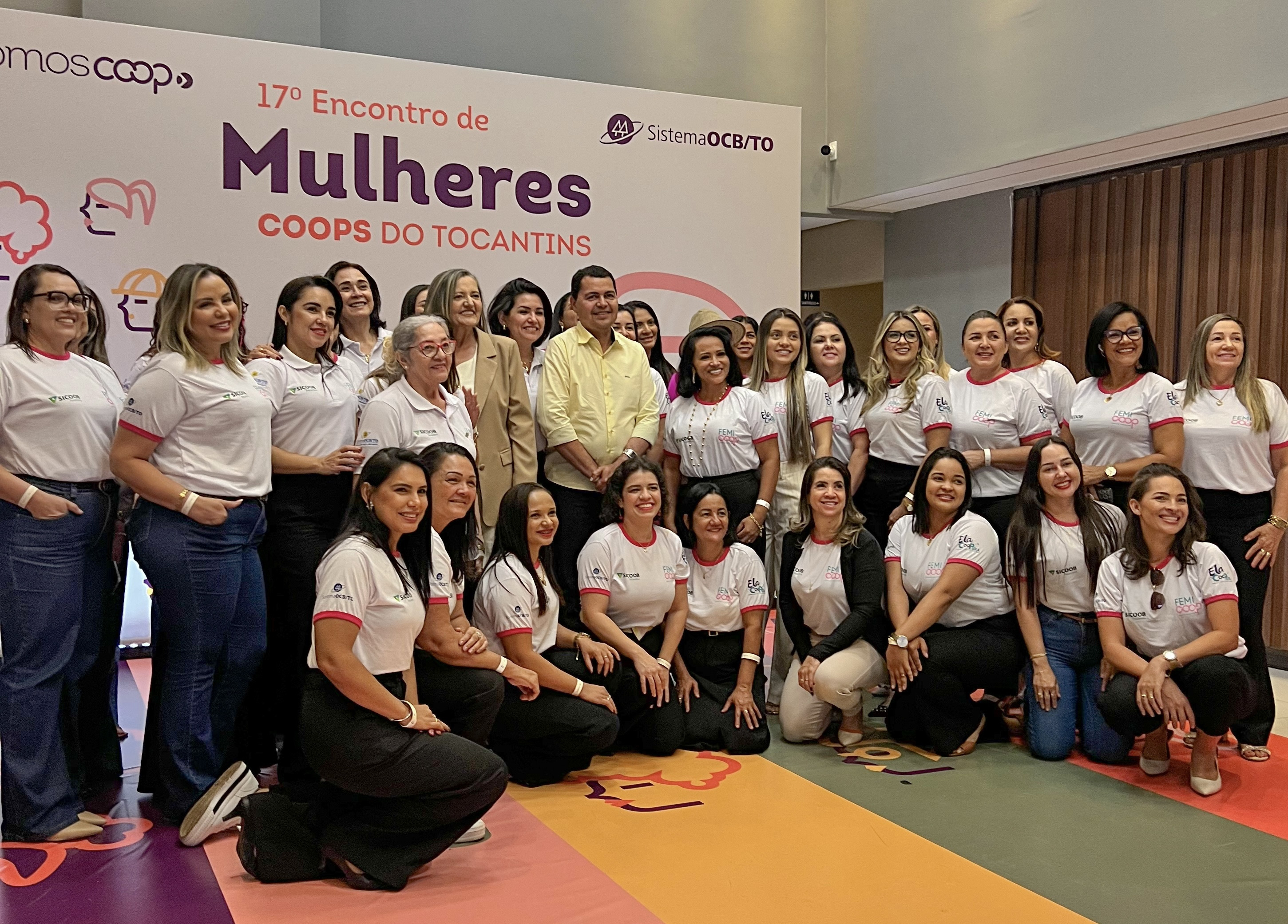 A imagem mostra um grupo grande de mulheres, com alguns homens misturados, posando para uma foto em um evento. Elas estão em frente a um banner branco com o título &quot;17º Encontro de Mulheres COOPS DO TOCANTINS&quot;. Muitas das mulheres usam camisetas brancas com detalhes em rosa e logotipos, e algumas usam calças jeans ou pretas. O ambiente parece ser um salão de eventos com iluminação clara. O chão tem um design colorido com formas geométricas. O tom geral da foto é de celebração e união.
