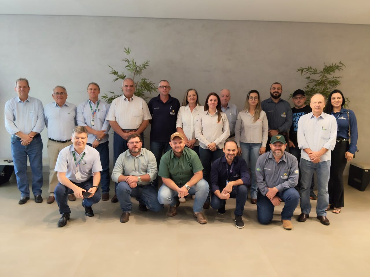 A imagem mostra um grupo de pessoas posando para uma foto em um ambiente interno. Há homens e mulheres, a maioria vestindo camisas claras e calças jeans. Eles estão organizados em duas fileiras, alguns em pé e outros agachados. O fundo é uma parede clara e há plantas decorativas ao redor.