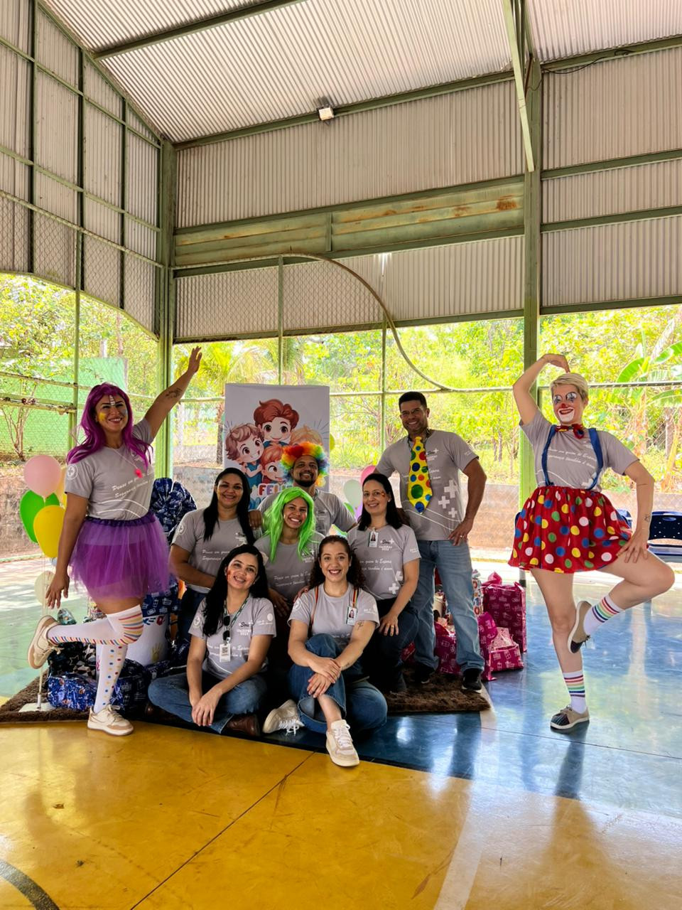 Esta é uma foto de um grupo de nove pessoas em um espaço coberto. Duas pessoas estão fantasiadas de palhaços, com roupas coloridas e perucas. As outras vestem camisetas cinza. Ao fundo, há um cartaz com desenhos de crianças e balões coloridos. Todos estão sorrindo e parecem felizes.